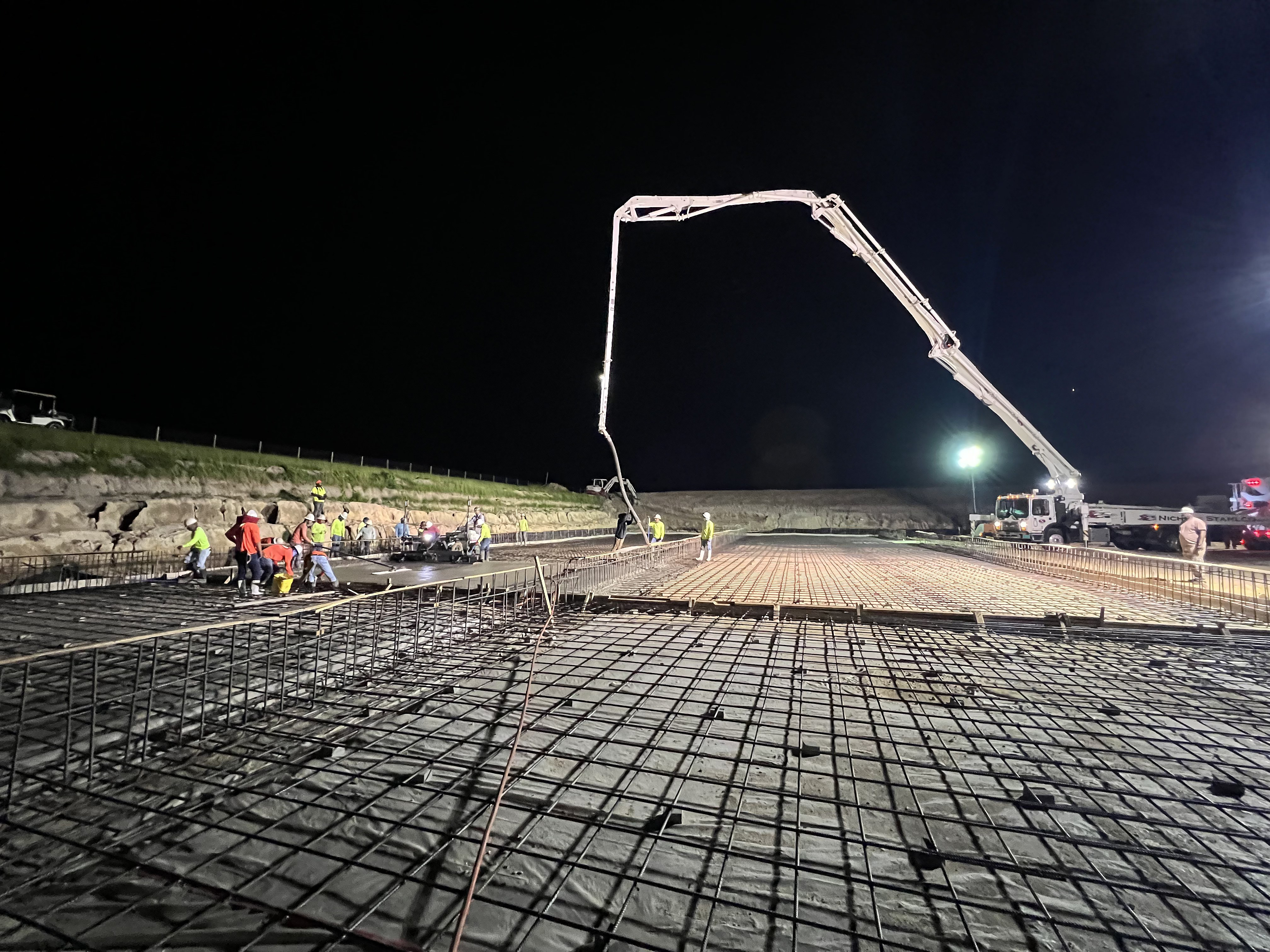Concrete pump boom placing a slab over a rebar grid at night with crew in hi-vis